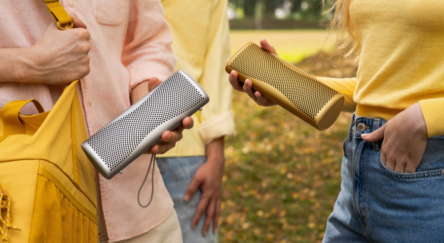 Zwei Personen halten tragbare Bluetooth-Lautsprecher im Freien, einen in Silber und einen in Gold, während eines ungezwungenen Treffens im Park.