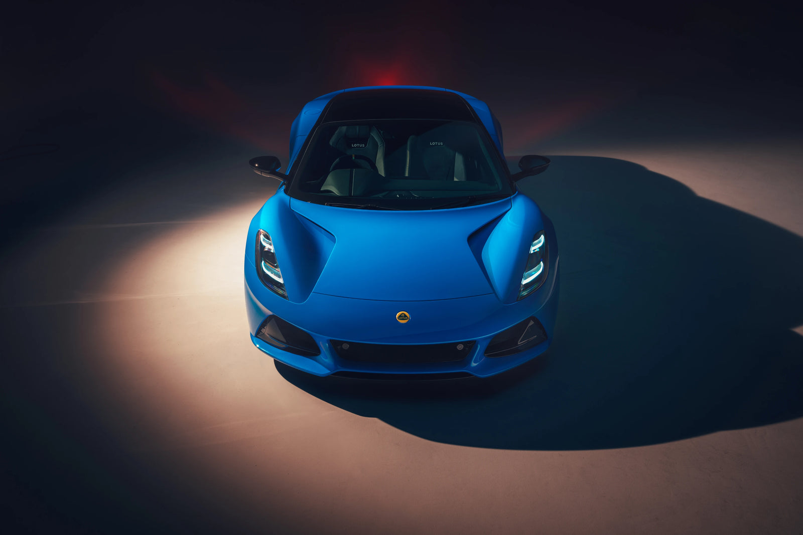 A bright blue Lotus Emira sports car photographed from above in a dark studio setting with dramatic spotlighting and a visible shadow.